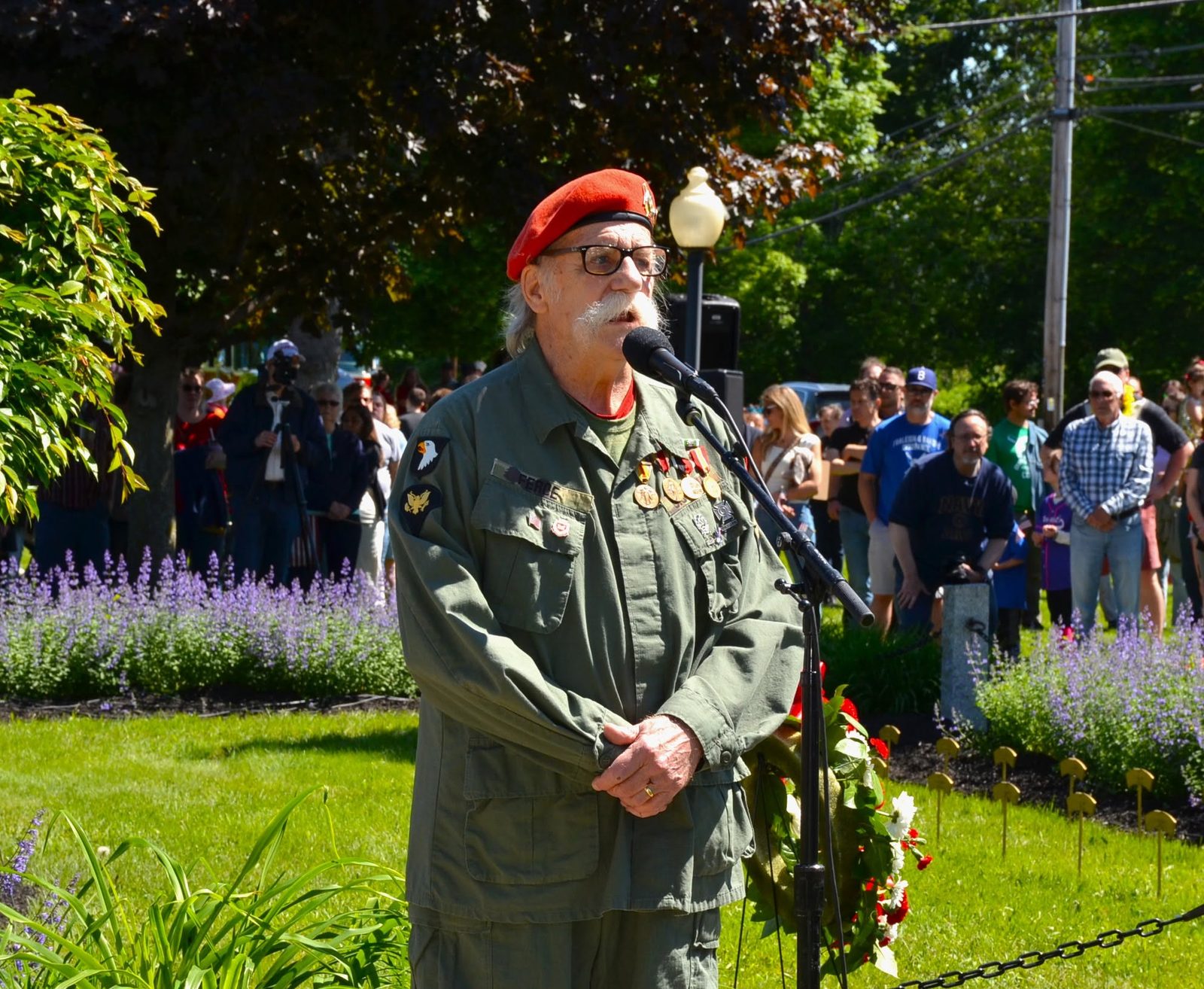 A Veteran in a red beret and field jacket speaking at a memorial ceremony as a crowd looks on